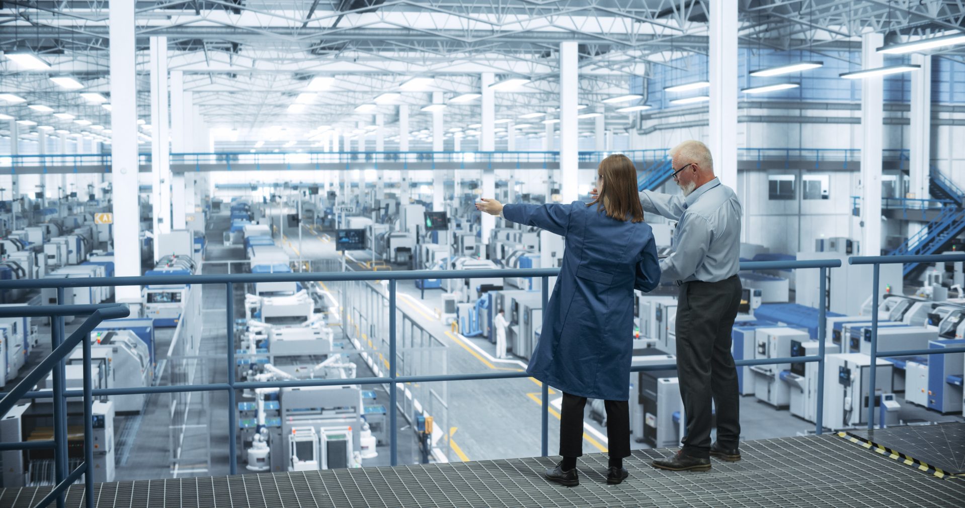 Experienced Male and Female Engineers Standing on a Platform with Their Back to Camera, Using Laptop Computer and Discussing Production at a Modern AI Automated Electronics Manufacture