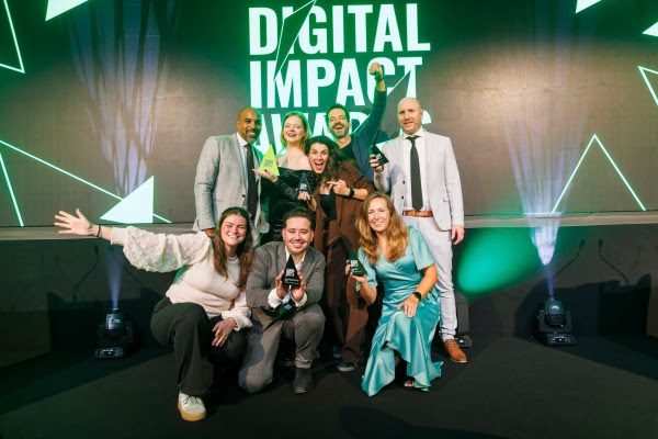 Een diverse groep van negen mensen staat op een podium. De mensen zijn in feestelijke kleding en poseren voor de foto met hun prijzen. Op de achtergrond is een groot scherm te zien met de woorden 'Digital Impact Awards' in het groen.
