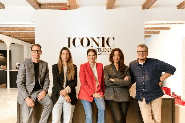 Een zakelijke portretfoto van vijf lachende mensen, die zij aan zij poseren in een modern kantoor. De groep bestaat uit twee vrouwen en drie mannen. Achter hen staat in grote gouden letters 'ICONIC STUDIOS'.